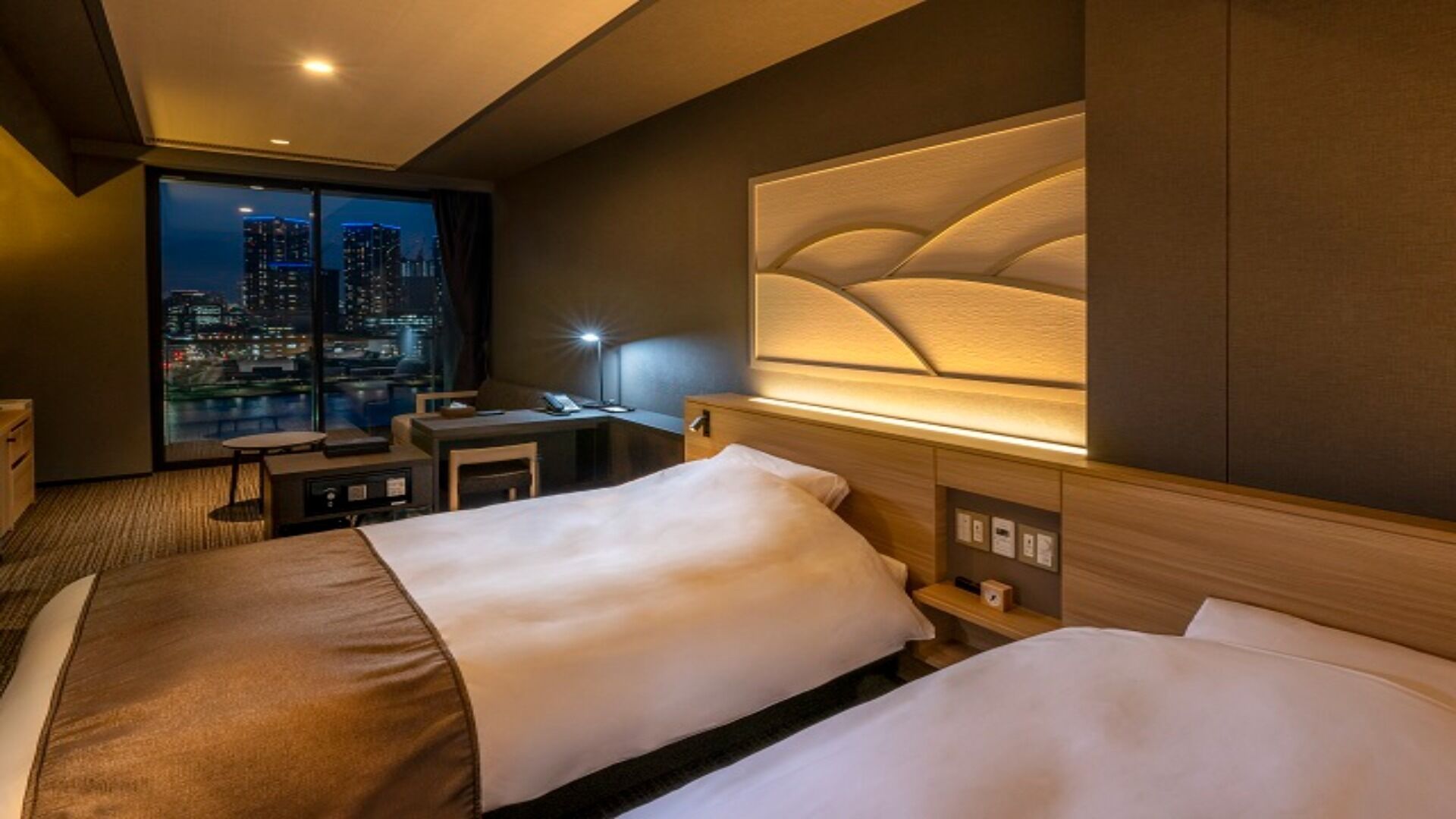 A hotel room with two beds, a desk, a chair, and a large window overlooking a cityscape at night.
