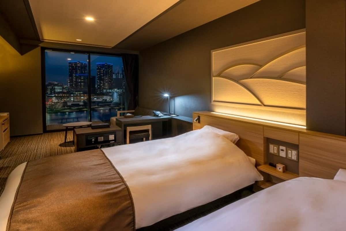 Habitación de hotel con dos camas, un escritorio, una silla y un ventanal grande con vistas a un paisaje urbano nocturno.