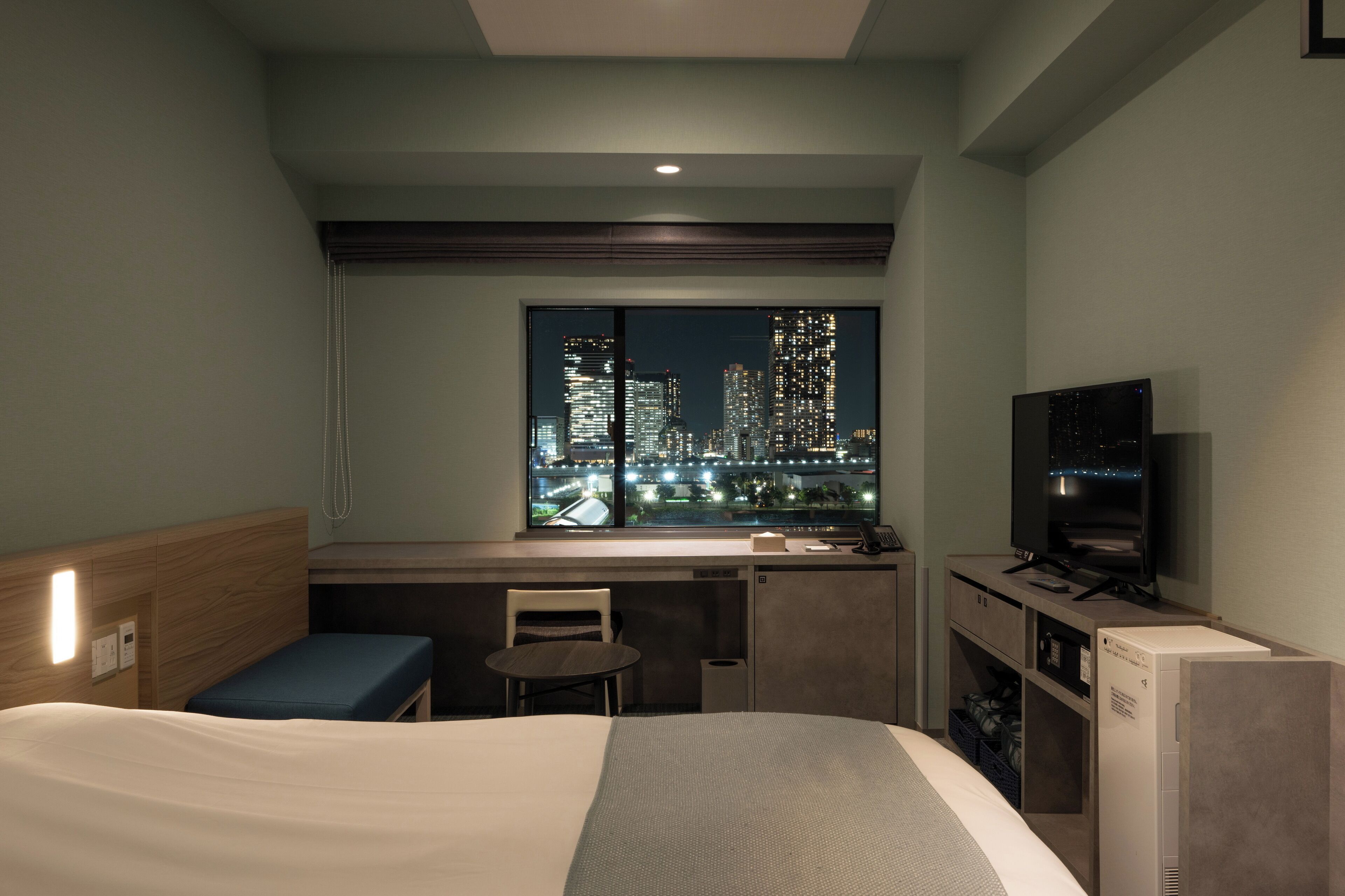 Ein Hotelzimmer mit Bett, Schreibtisch, Stuhl, Fernseher und einem Fenster mit Blick auf die Stadt.
