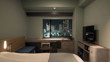 Ein Hotelzimmer mit Bett, Schreibtisch, Stuhl, Fernseher und einem Fenster mit Blick auf die Stadt.