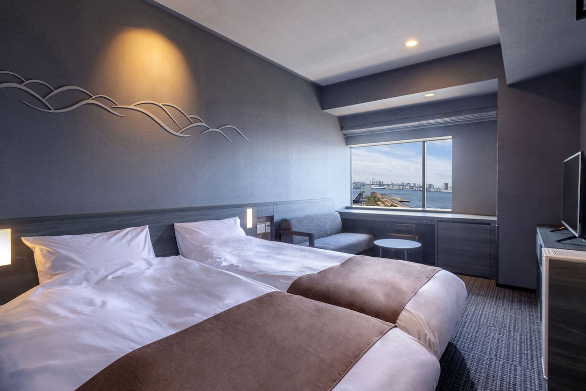 Una habitación de hotel moderna con una cama grande, dos sillones, una mesita y vistas a un paisaje urbano a través de un ventanal amplio.