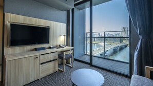 Une chambre d’hôtel moderne dotée d’une grande télévision, d’un bureau, d’une chaise et offrant une vue sur le paysage urbain grâce à de grandes fenêtres.