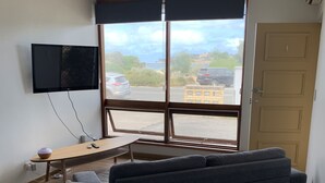 A room with a large window, a flat-screen TV mounted on the wall, a sofa, a dining table with chairs, and a small table with a lamp.