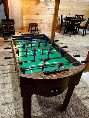 Taula de futbolí de fusta en una habitació amb terres i parets de fusta, un petit tamboret de fusta i una taula amb caires.