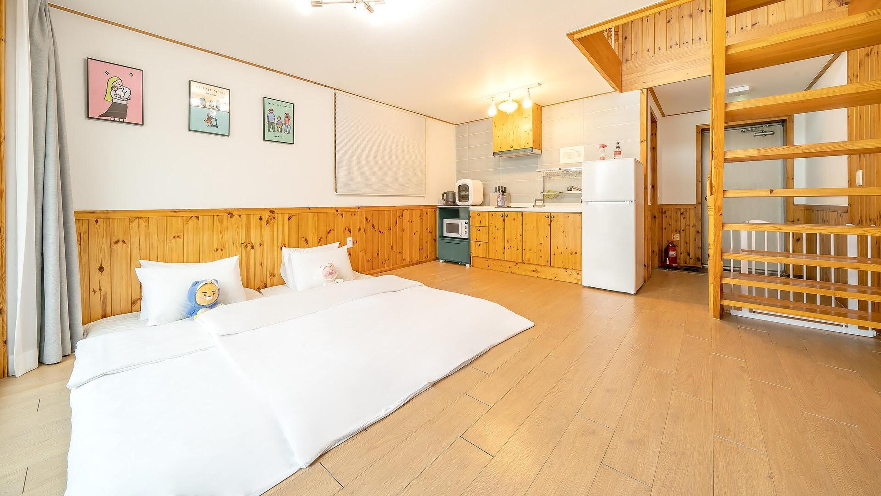 A bedroom with a wooden headboard, a bed with white bedding, a small kitchenette with a microwave and refrigerator, and a wooden staircase.