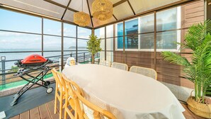 A sunlit outdoor dining area with a view of the sea, a barbecue grill, and wicker furniture. - Yeosu Hi Mom Kids Family Pension Black (Yeosu)