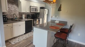 Eine moderne Küche mit weißen Schränken, einer eingebauten Mikrowelle, einem Herd und einem Kühlschrank. Es gibt eine Bar mit zwei Hockern und eine kleine Kochinsel mit einem Spülbecken.