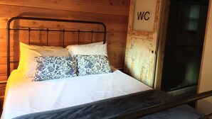 A wooden-paneled room with a bed featuring patterned pillows and a metal headboard. A door with "WC" is visible to the right.