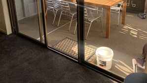 Una porta in vetro conduce a un patio esterno con un tavolo e sedie in legno, un barbecue e un secchio con l'etichetta "BUY RIGHT".
