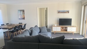 Un soggiorno moderno con un divano angolare grigio, un tavolo da pranzo in legno con sedie, una TV su un mobile in legno e un quadro appeso al muro.