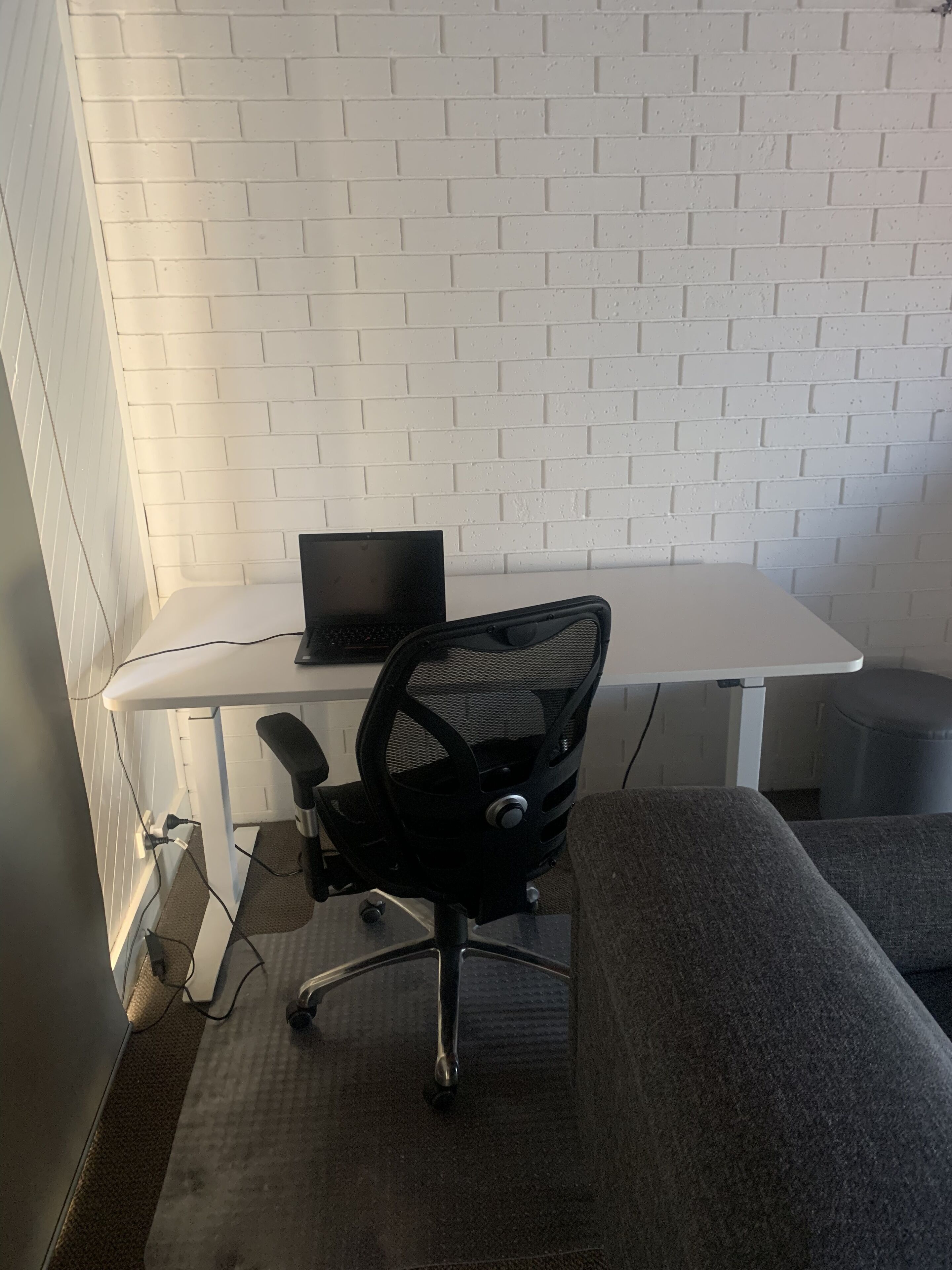 Un'area lavoro con una parete in mattoni bianchi, una sedia da ufficio nera, una scrivania con un portatile e un divano grigio.