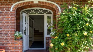 A brick building entrance with a white door, a small porch, and a potted plant. - Whg. Nordstrand - House on the dike (Utersum)