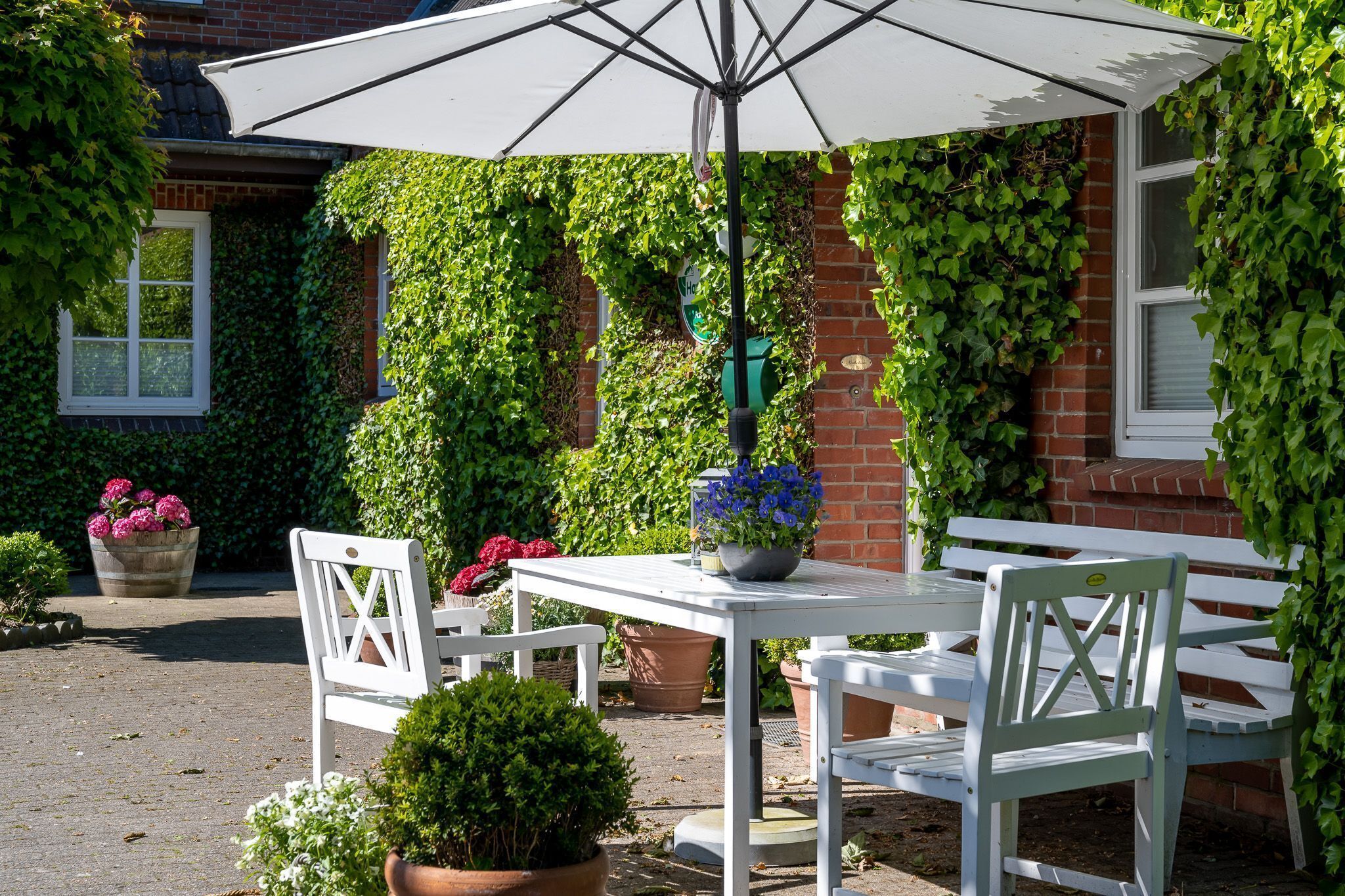 Eine Terrasse im Freien mit einem weißen Tisch und einer Bank, umgeben von Topfpflanzen und einer mit Efeu bewachsenen Backsteinmauer.