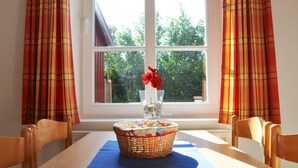 A dining table with a wicker basket, a vase with red flowers, and a window with plaid curtains. - Whg. Nordstrand - House on the dike (Utersum)