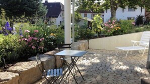 Une terrasse extérieure avec une table et deux chaises, entourée d’un jardin où poussent différentes plantes et fleurs.