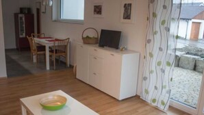 Une pièce comprenant une table à manger, des chaises, un meuble blanc avec un téléviseur et une fenêtre.
