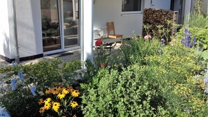 Un patio abrité avec une table et des chaises, une pile de bois de chauffage et diverses plantes à fleurs.