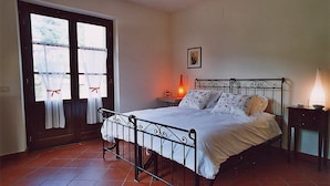 Un dormitorio con estructura de cama metálica, mesita de noche con lámpara y ventana con cortinas.