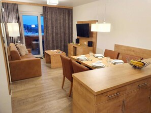 A modern hotel room with a wooden dining table set for four, a sofa, a TV mounted on the wall, and a balcony with curtains.