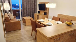 Una camera d'albergo moderna con un tavolo da pranzo in legno apparecchiato per quattro persone, un divano, una TV a parete e un balcone con tende.
