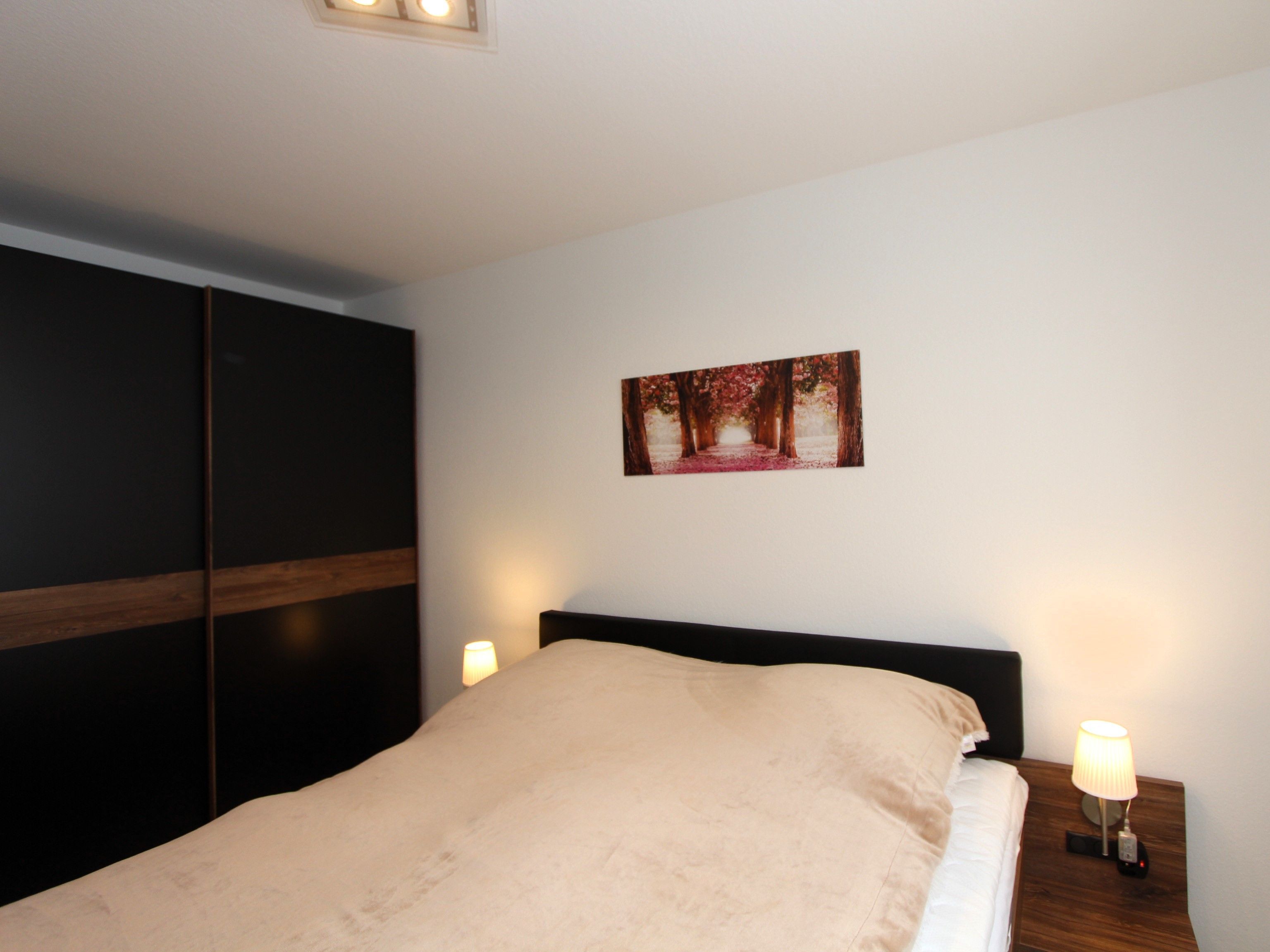 A bed with a beige blanket, a wooden headboard, a nightstand with a lamp, and a framed picture on the wall.