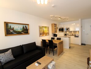 A modern living room with a black sofa, a wooden coffee table, and a dining area with a wooden table and chairs. A kitchen with white cabinets and stainless steel appliances is visible in the background.