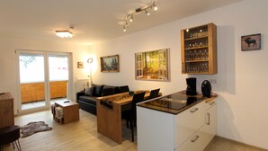A modern living room with a sofa, coffee table, and dining table. There is a kitchen area with a coffee maker and cabinets. The room has a large window and a painting on the wall. - GRA-115 - South-facing vacation apartment with whirlpool - Residence Grafenmatt 1.2 (Feldberg (Schwarzwald))