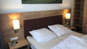 A hotel room with a bed, two bedside lamps, a nightstand with a plant, a wooden headboard, and a large landscape painting on the wall.