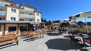 A terrace with wooden benches and tables, a building with balconies, and outdoor umbrellas.