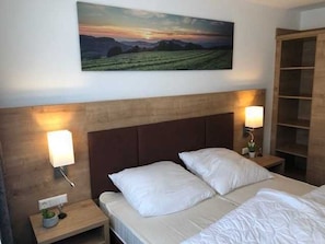 Une chambre d’hôtel avec un lit, deux lampes de chevet, une table de chevet ornée d’une plante, une tête de lit en bois et un grand tableau représentant un paysage accroché au mur.