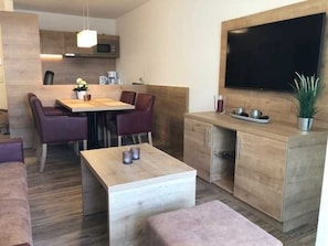 Une cuisine moderne avec des meubles en bois, un coin repas avec une table et des chaises, un téléviseur fixé au mur et une petite plante en pot.