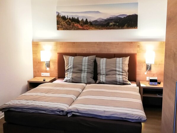 A double bed with striped bedding, two bedside lamps, a nightstand, and a wall-mounted painting of a landscape.
