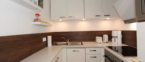 Cozinha compacta com armários brancos, pia de aço inoxidável, forno embutido e cafeteira.