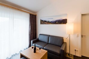A modern living room with a sofa, a coffee table with wine glasses and a bottle, a lamp, and a painting of a landscape on the wall.