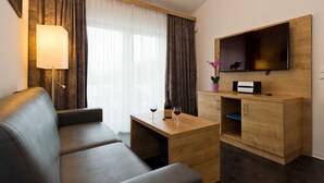 Une chambre d’hôtel moderne équipée d’un canapé, d’une table d’appoint en bois sur laquelle se trouvent un verre et une bouteille de vin, d’un téléviseur à écran plat fixé à un mur en bois, et d’une petite armoire contenant une imprimante.