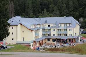 A multi-story hotel with balconies, a covered outdoor seating area, and a view of a forested area.