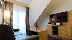 A modern hotel room with a sofa, a small wooden table, a flat-screen TV, a potted plant, and a large window with curtains.
