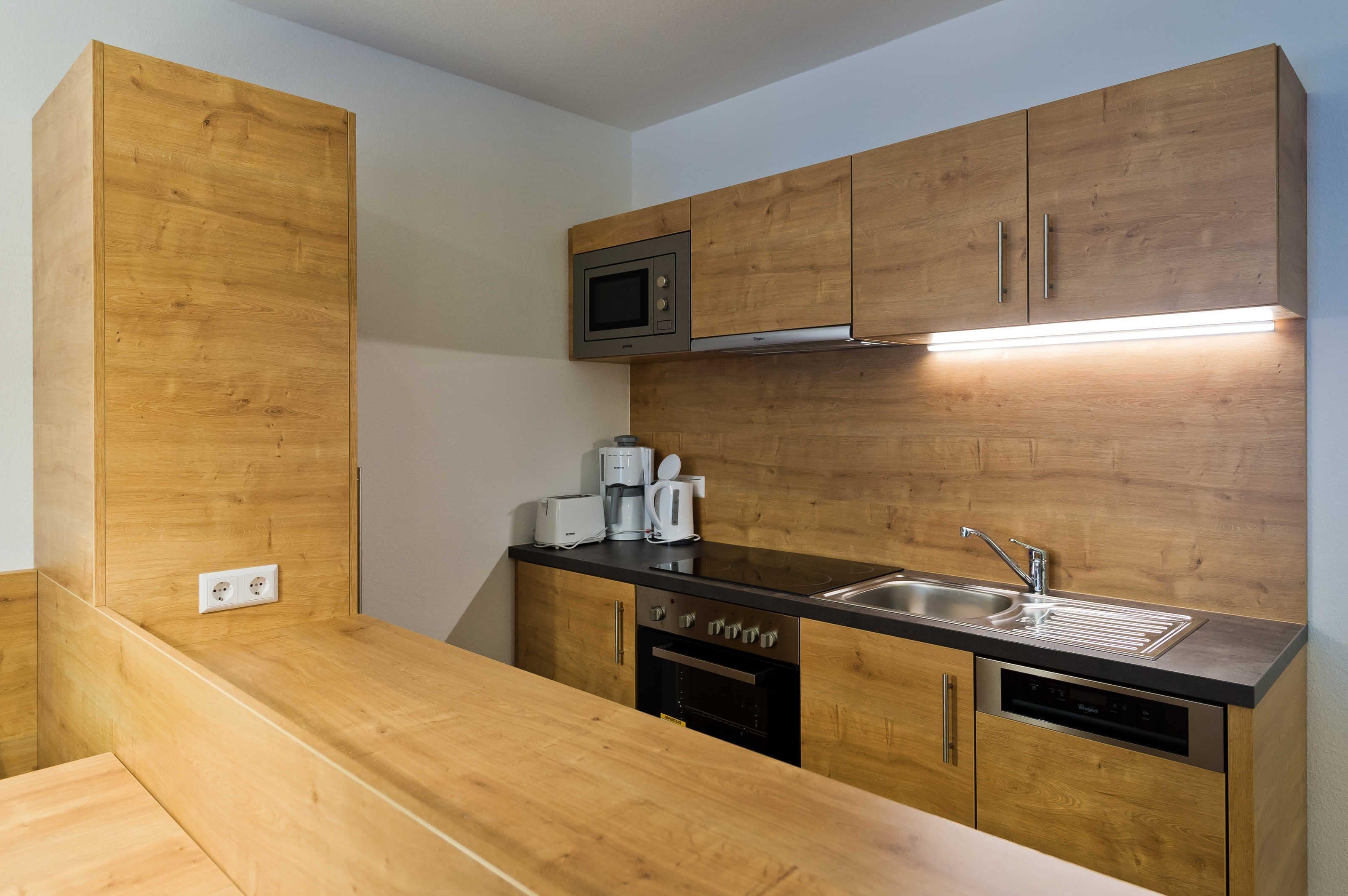 A compact kitchen with wooden cabinets, a microwave, a coffee maker, a toaster, and a dishwasher.
