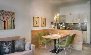 Een moderne keuken met witte kastjes, een eethoek met een houten tafel en groene stoelen, en een schilderij van een boom aan de muur.