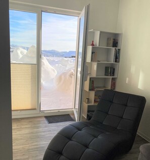 Une pièce dotée d’une porte-fenêtre coulissante donnant sur un paysage enneigé, d’un fauteuil sombre et d’une bibliothèque remplie de livres et d’objets décoratifs.