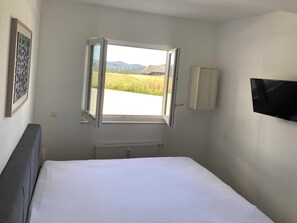 Un dormitorio con cama, ventana con vistas abiertas, radiador, televisor montado en la pared y un cuadro enmarcado.