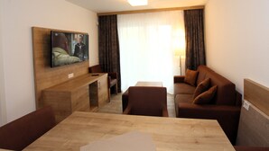 Une chambre d’hôtel avec une table à manger en bois, un canapé marron, un téléviseur fixé au mur et une fenêtre avec des rideaux.