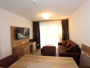 Ein Hotelzimmer mit einem Holztisch, einem braunen Sofa, einem an der Wand befestigten Fernseher und einem Fenster mit Vorhängen.