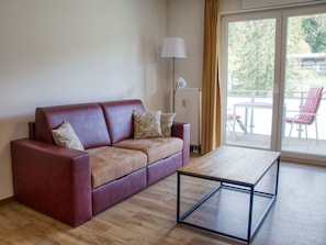 Un sofá de cuero rojo con cojines, una mesa de centro de madera, una lámpara de pie y un ventanal corredizo que da a un área exterior con sillas.