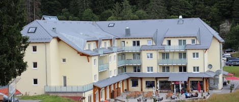 A multi-story hotel with balconies, a covered outdoor seating area, and a view of a forested area.