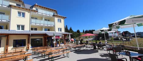 A terrace with wooden benches and tables, a building with balconies, and outdoor umbrellas.