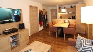 A modern living room with a flat-screen TV, wooden flooring, a dining area with a table and chairs, and a kitchen area with cabinets and a hanging light fixture. - GRA-217 - Comfortable apartment with whirlpool - Residence Grafenmatt 2.2 (Feldberg (Schwarzwald))
