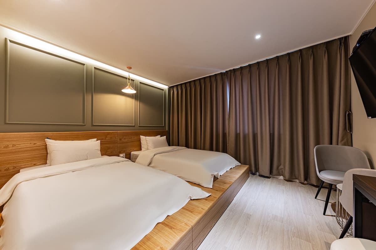 A hotel room with two beds, a wooden headboard, a desk with a chair, and a large window with curtains.