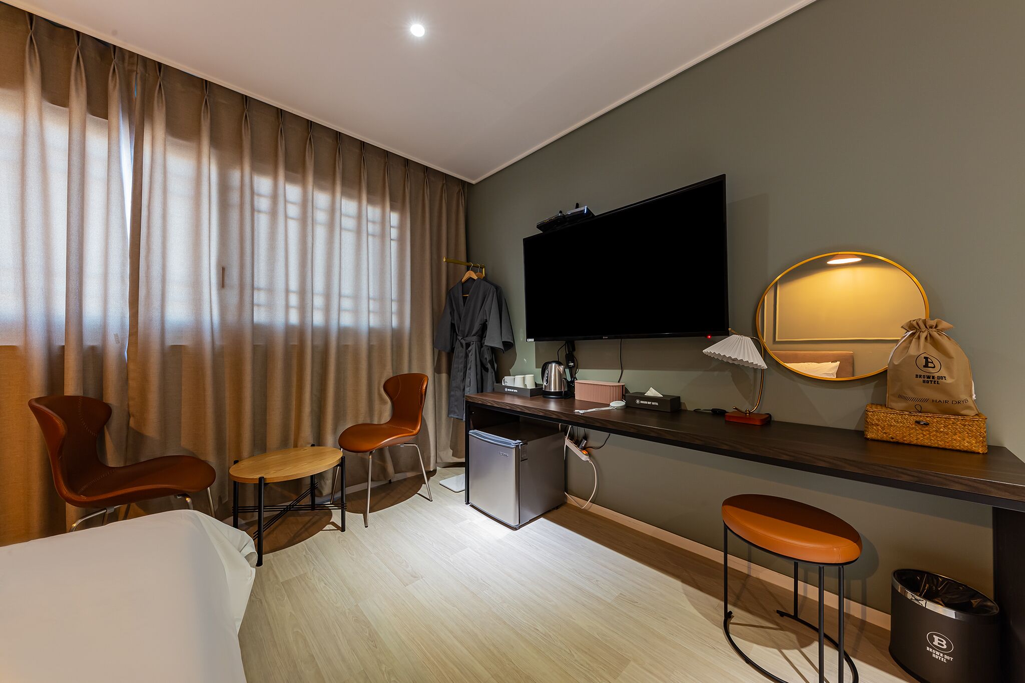 A modern hotel room with a bed, two chairs, a small table, a TV, a desk with a lamp, and a trash bin.
