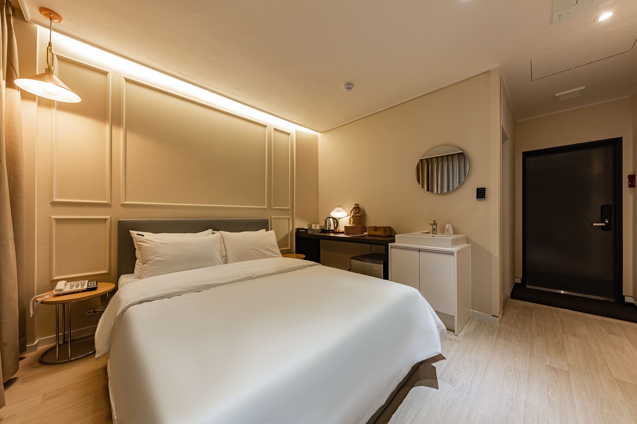 Een hotelkamer met een groot bed, nachtkastje, lamp en een klein bureau met spiegel en vaas.